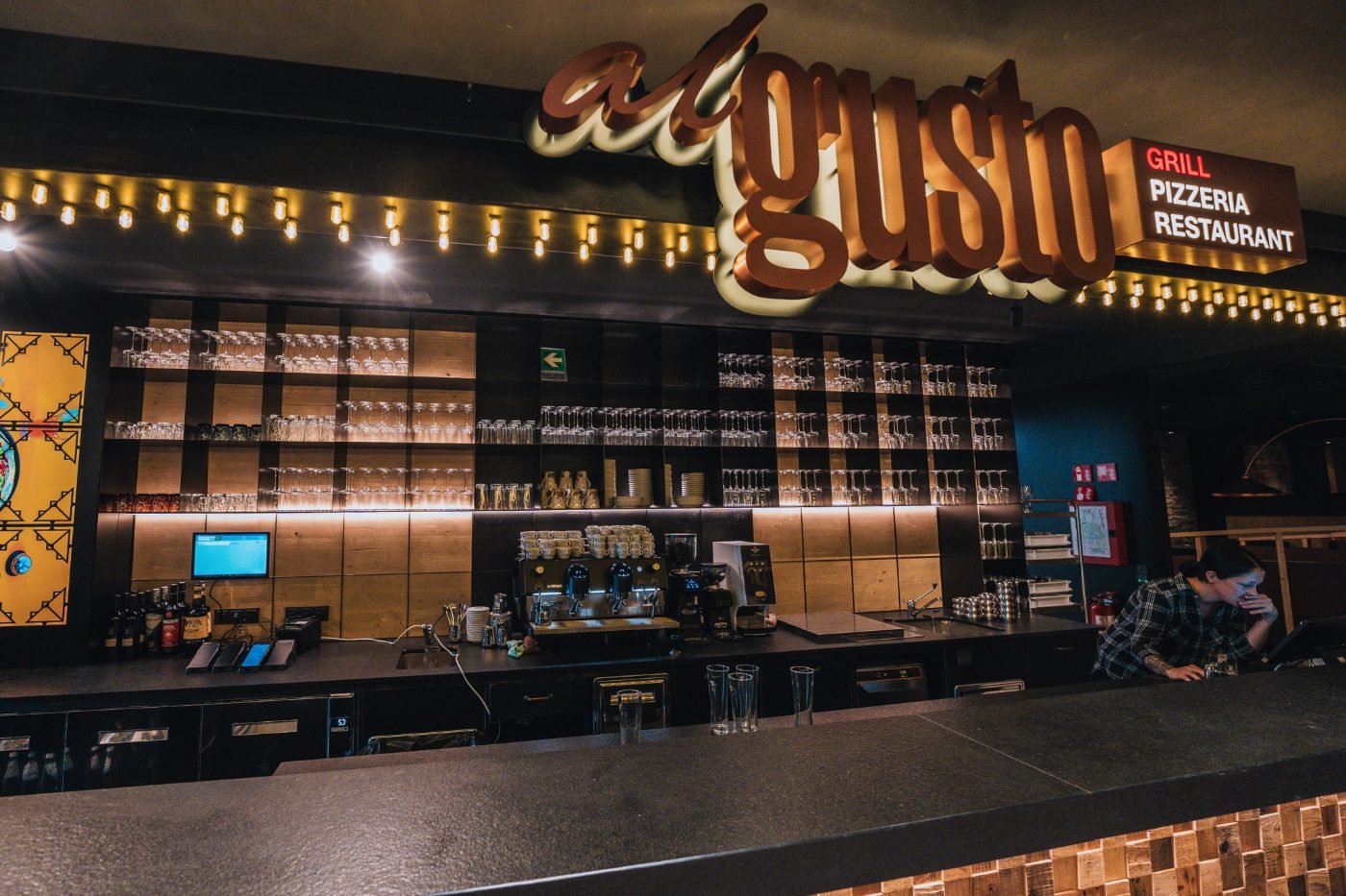 Die Bar im Al Gusto am Abend: beleuchteter Schriftzug, Glühbirnenketten, Regale voller Gläser